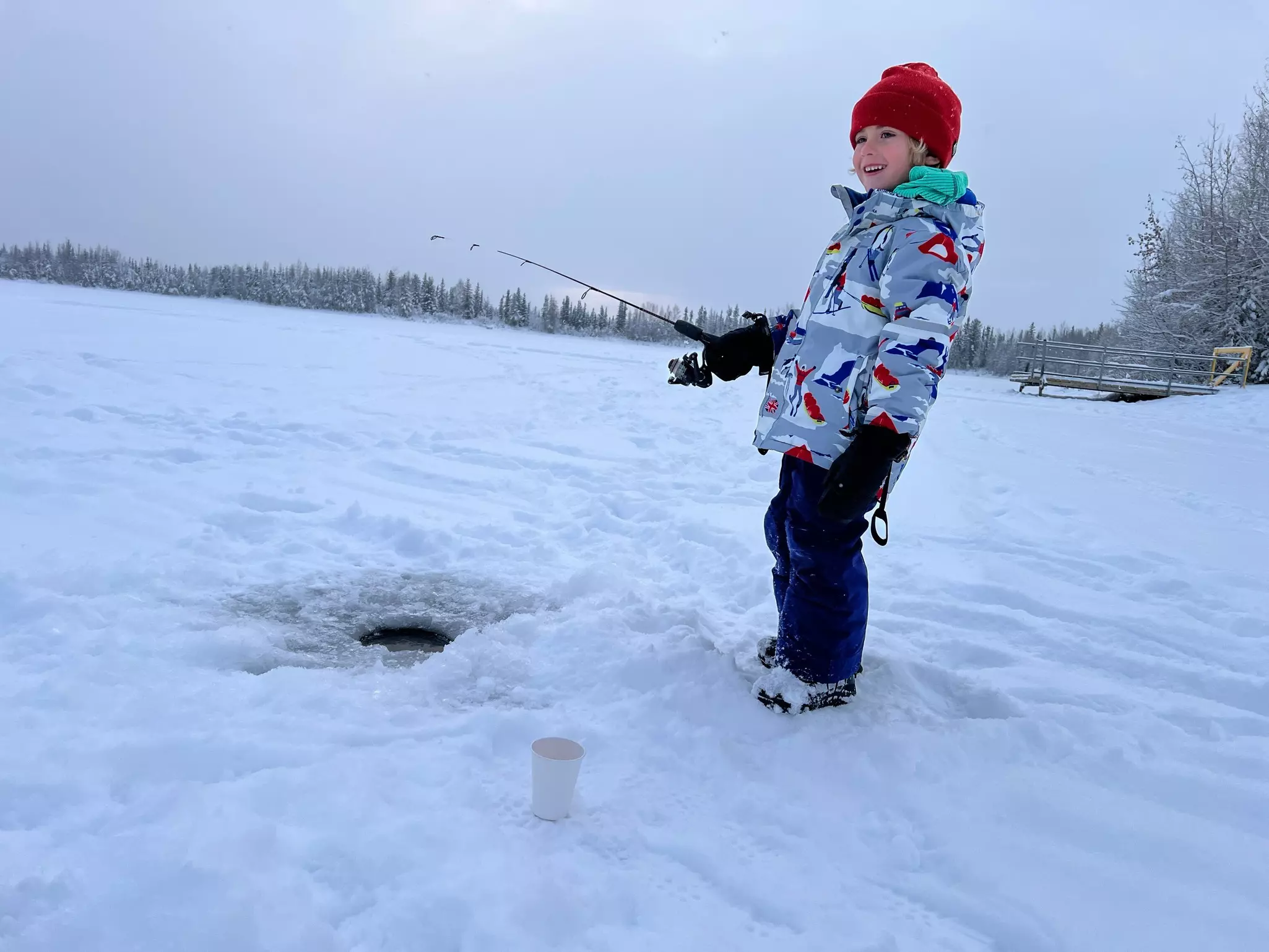 A boy wearing a red had, blue pants and a gray patterned jacket holds a fishing pole over a hole in snow-covered ice in Alaska.