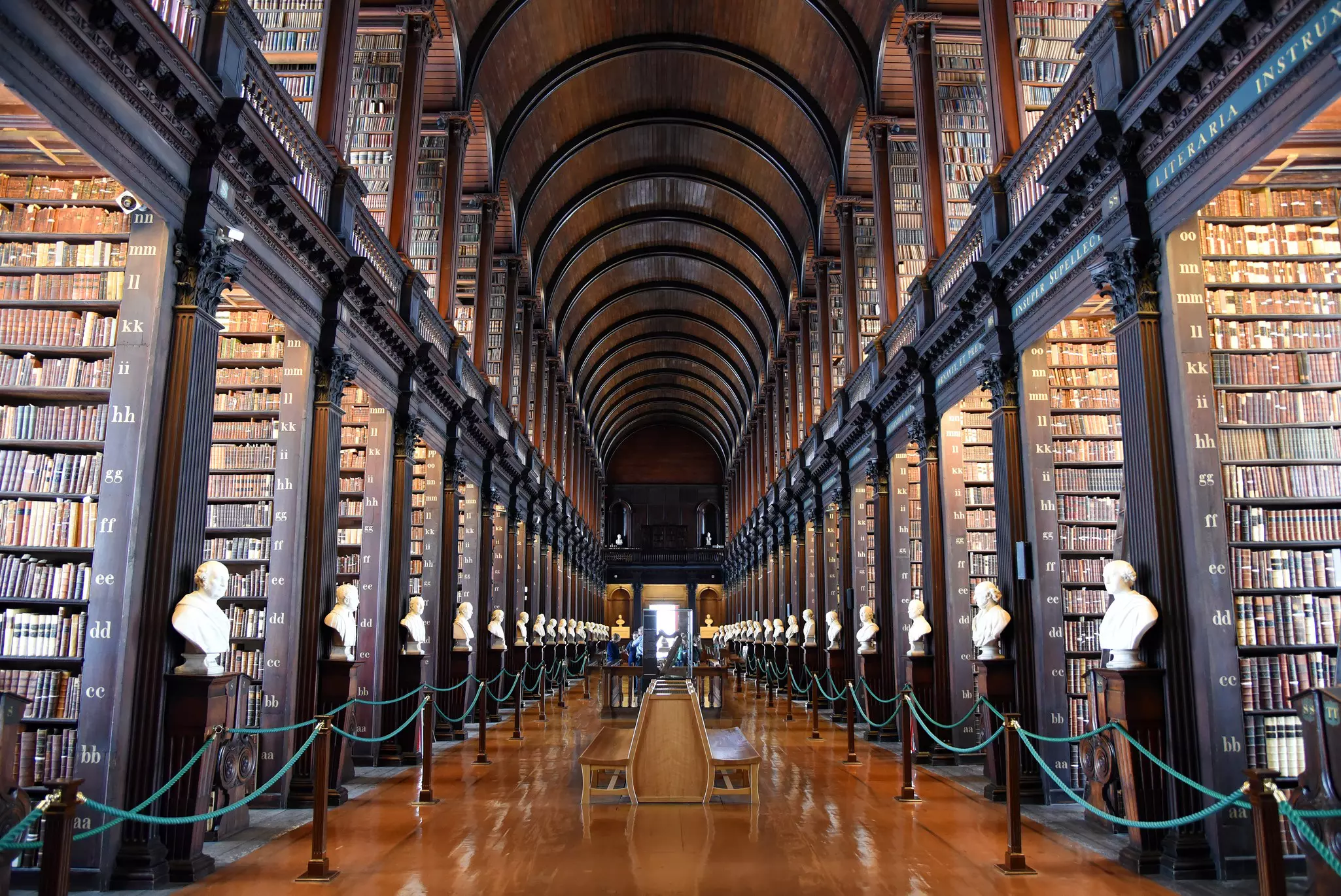 Two stories of floor-to-ceiling bookcases are lined up on either side of a central walkway with a wood floor and benches in the center; there are white busts at the end of each bookcase.