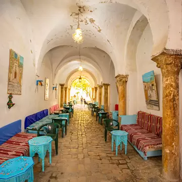 Long whitewashed vaulted passage with stone columns, cushioned benches and turquoise tables leading to a sunlit arch.