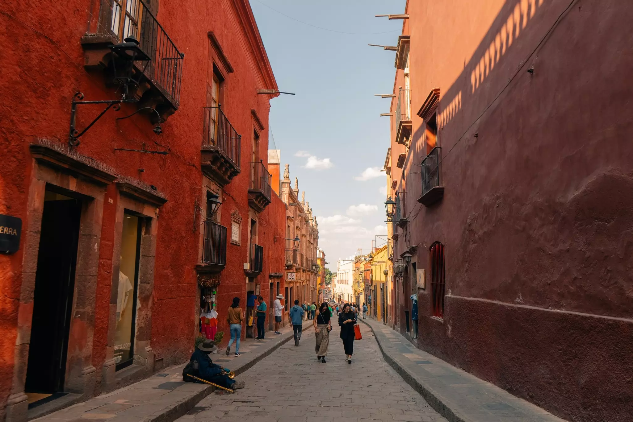People walk on a narrow street with buildings on either side; the street is in shade, the buildings are in warm reddish tones, and there is sunshine on the buildings in the far distance.