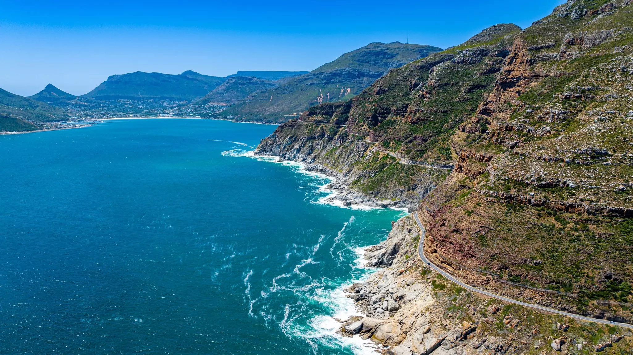 Aerial of bright blue ocean with narrow roadway along a rocky mountain with other mountains in the distance on a sunny day.