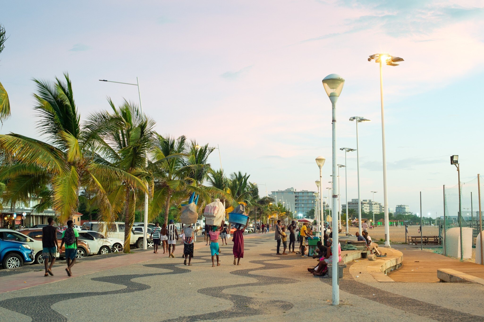 People walking a wave-patterned promenade lined with palms and streetlights on Ilha de Luanda, Angola, at dusk