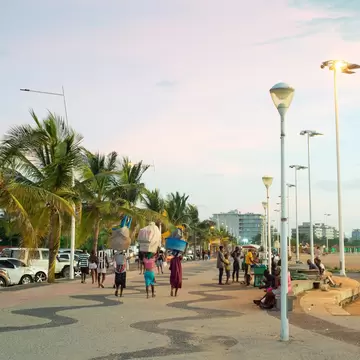 People walking a wave-patterned promenade lined with palms and streetlights on Ilha de Luanda, Angola, at dusk
