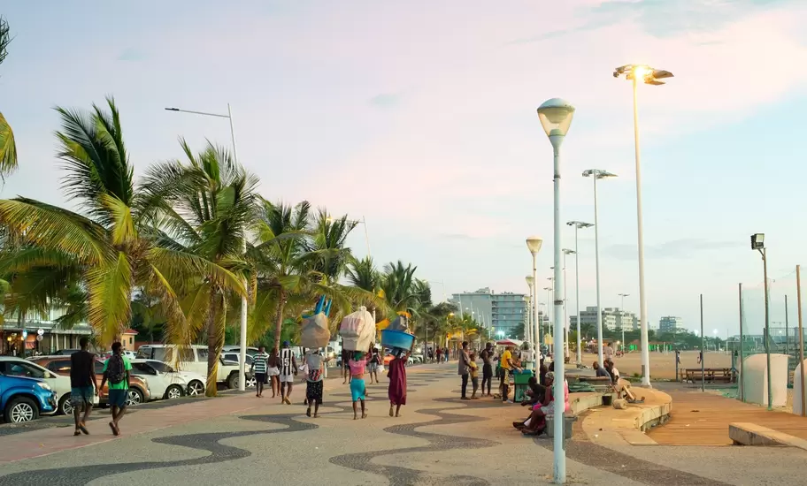 People walking a wave-patterned promenade lined with palms and streetlights on Ilha de Luanda, Angola, at dusk