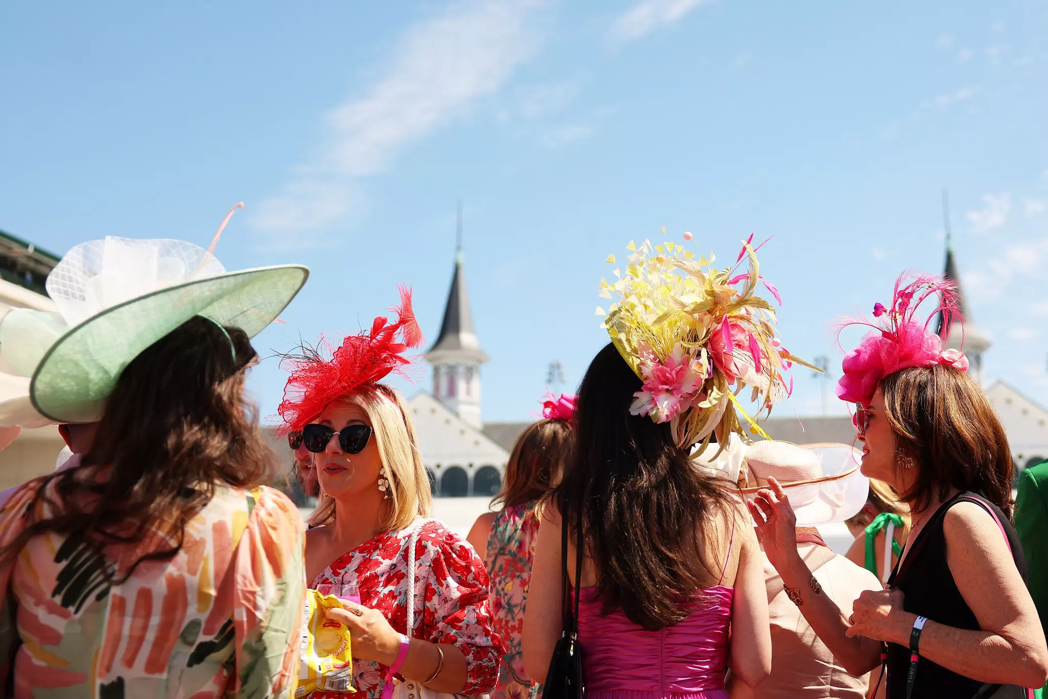 A group of women wears colorful dresses and elaborate hats with feathers at a horse racetrack.