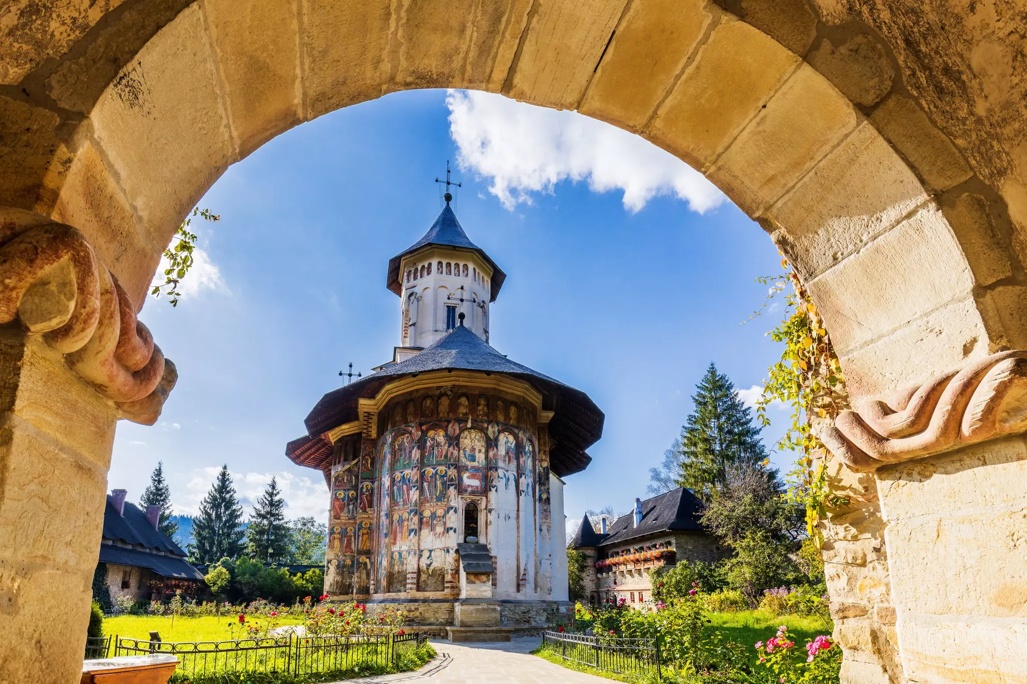 A monastery sits in a lush garden on a sunny day. The external walls are covered with frescoes that represent biblical scenes.
