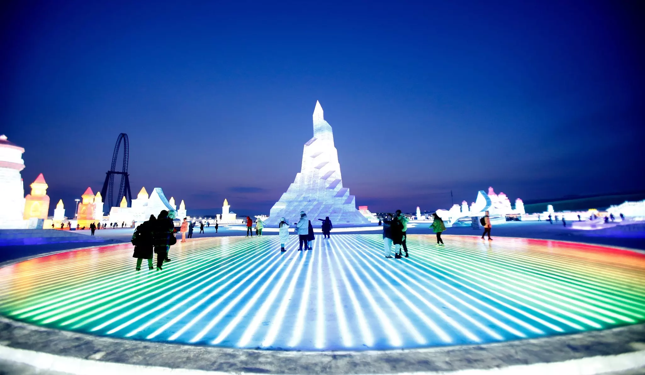 People walk over ice that is lit from below in an array of colors. A huge ice sculpture stands as tall as a building nearby