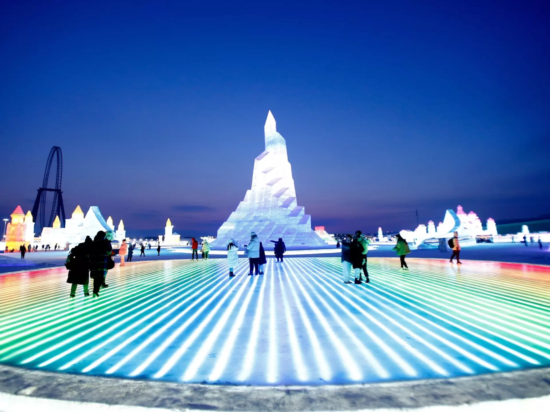People walk over ice that is lit from below in an array of colors. A huge ice sculpture stands as tall as a building nearby