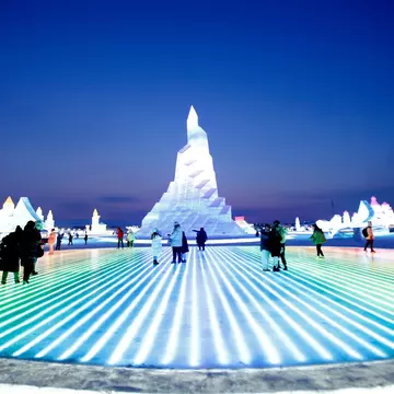 People walk over ice that is lit from below in an array of colors. A huge ice sculpture stands as tall as a building nearby