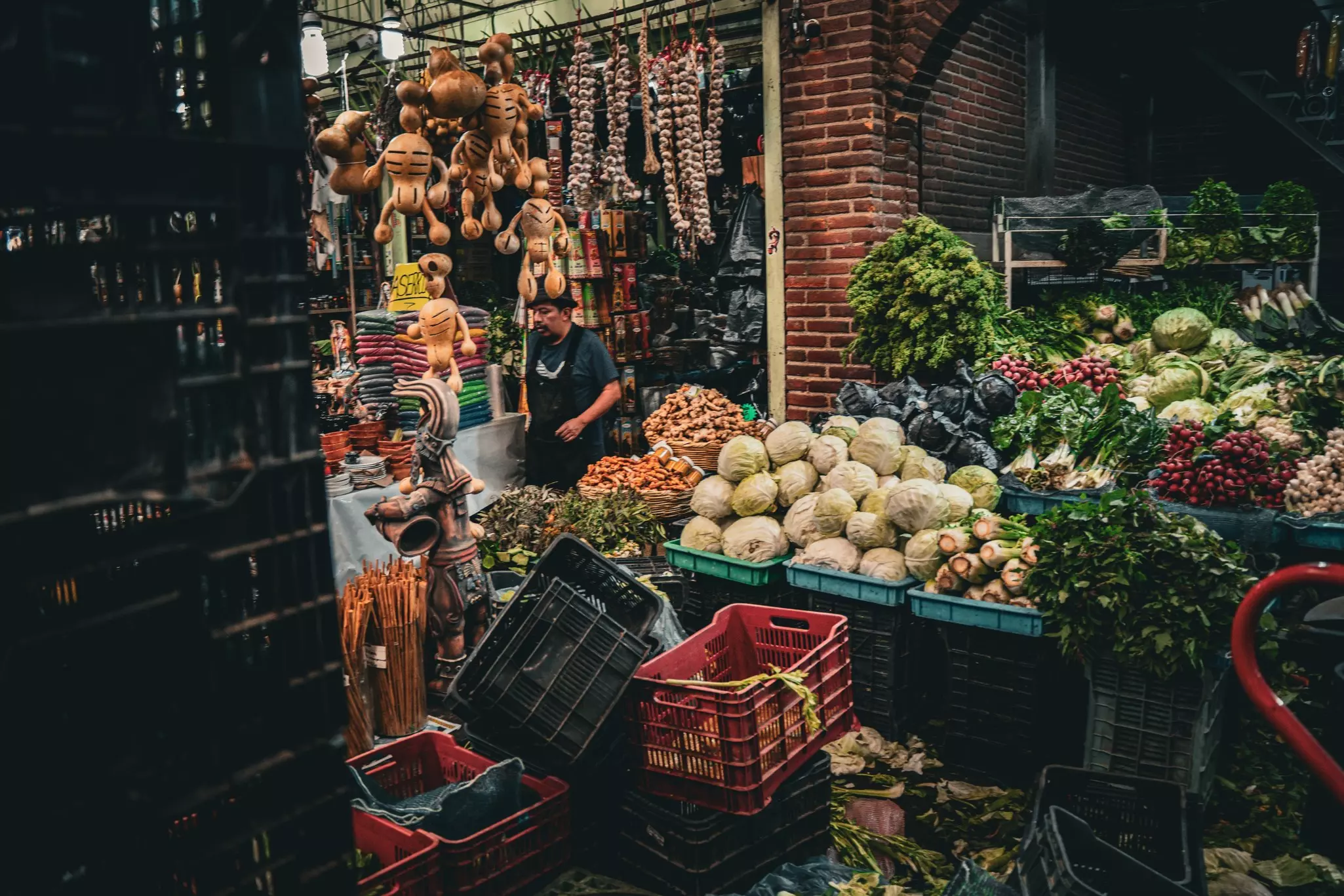 A vendor stands near stalls in a market fully loaded with fresh vegetables, herbs and handmade crafts. 
