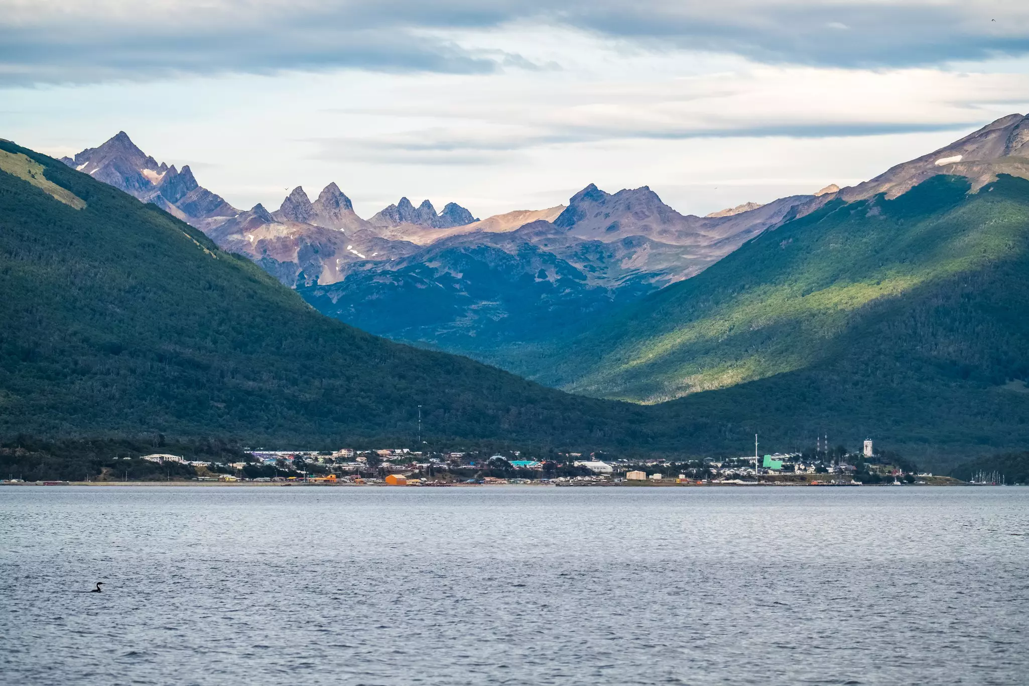 Looking across a sea at a small town nestled at the bottom of forested mountains with rocky mountains in the distance on a partly sunny day.