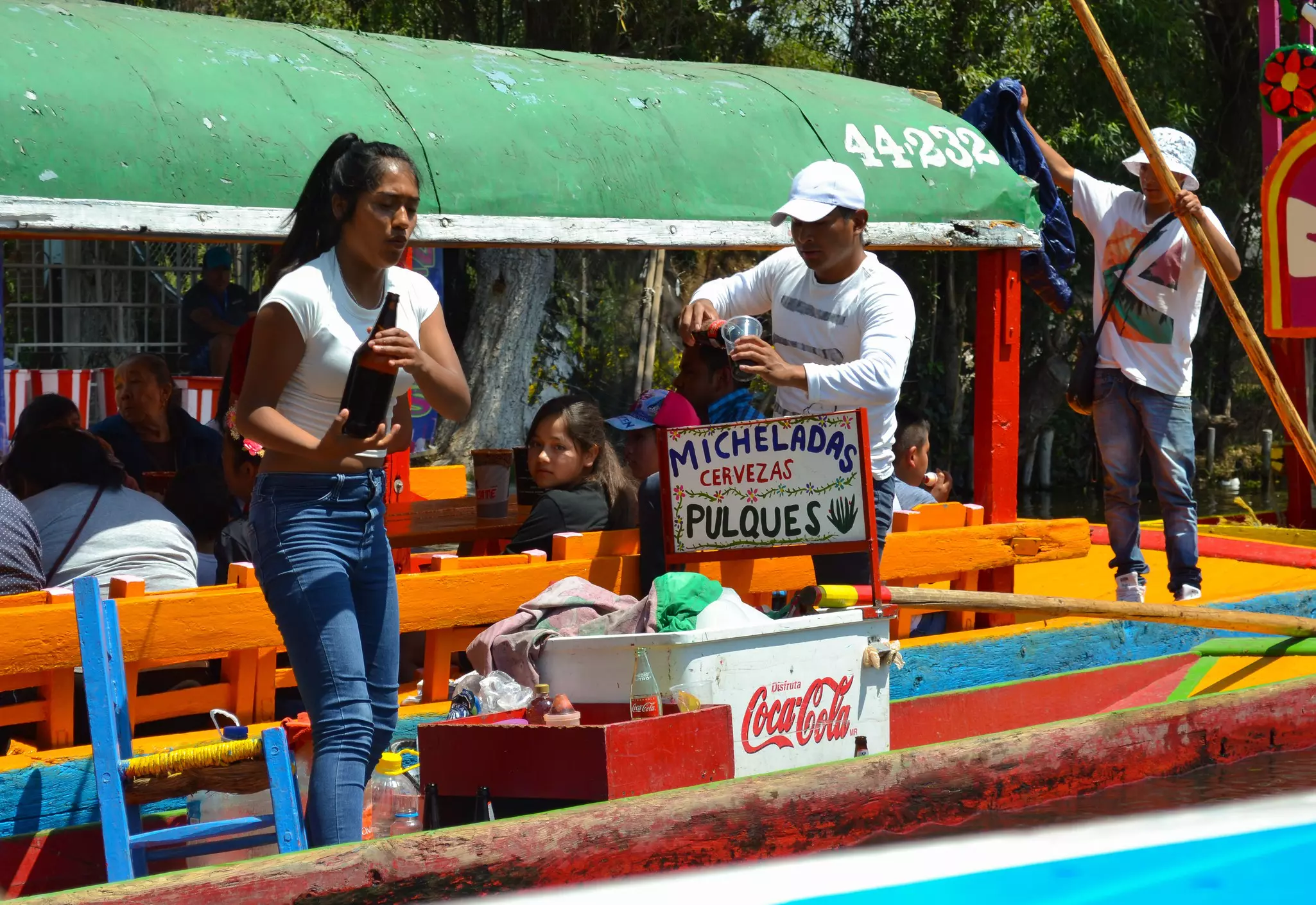 A person on a boat with an orange railing holds a large dark bottle; another person pours from another bottle into a plastic cup; a decorated sign by a cooler says, "Micheladas, cervezas, pulques."