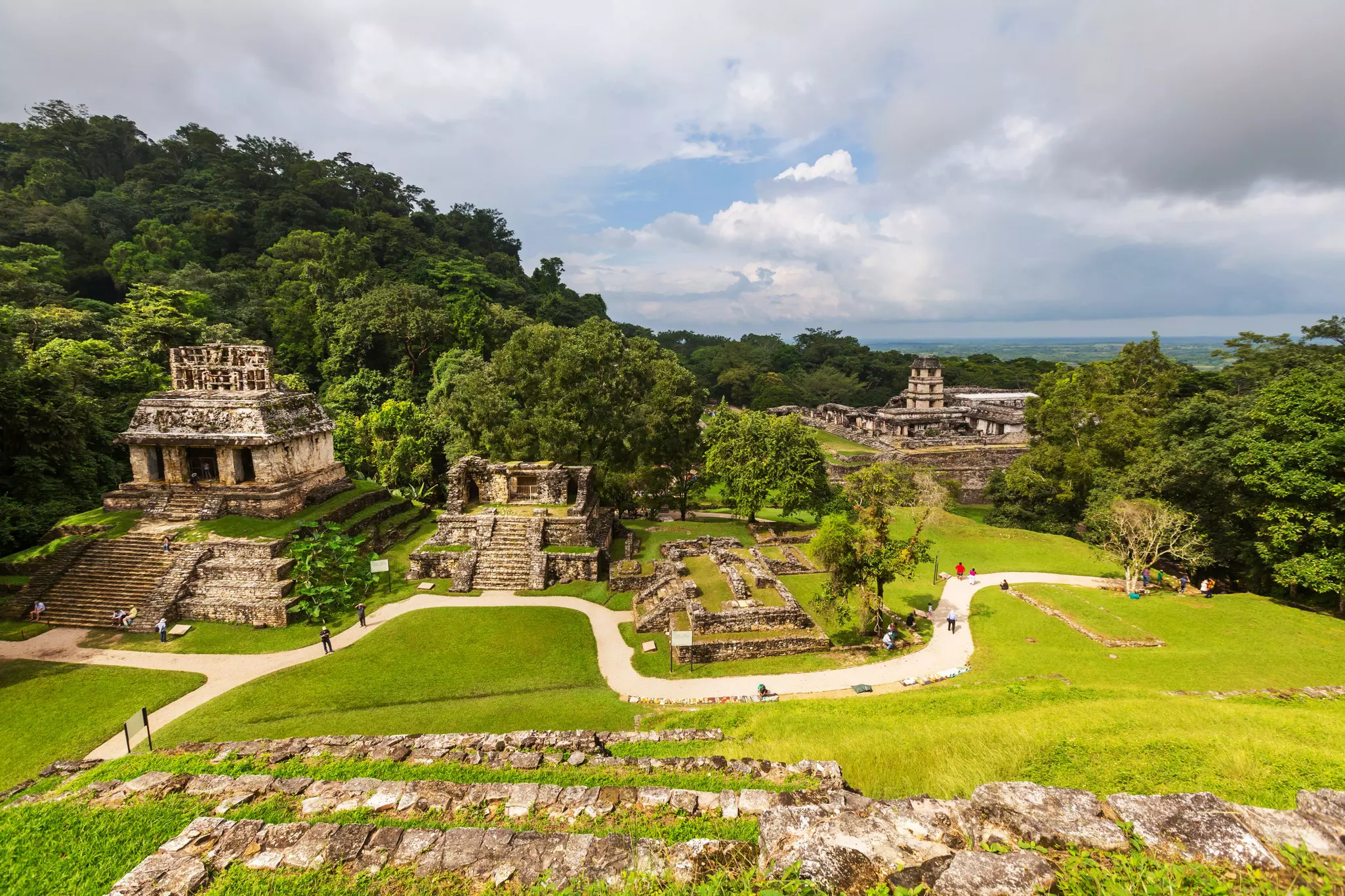 Tourists follow a path among ancient stone temples backed by jungle.