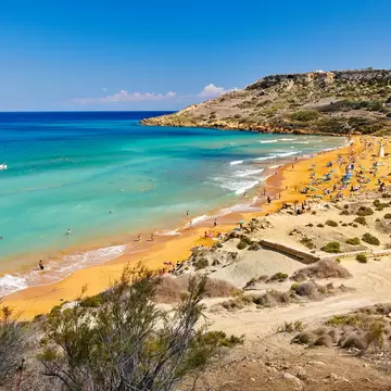 Long orange sand beach with bathers and umbrellas beside turquoise sea, scrub-covered slopes in the foreground.