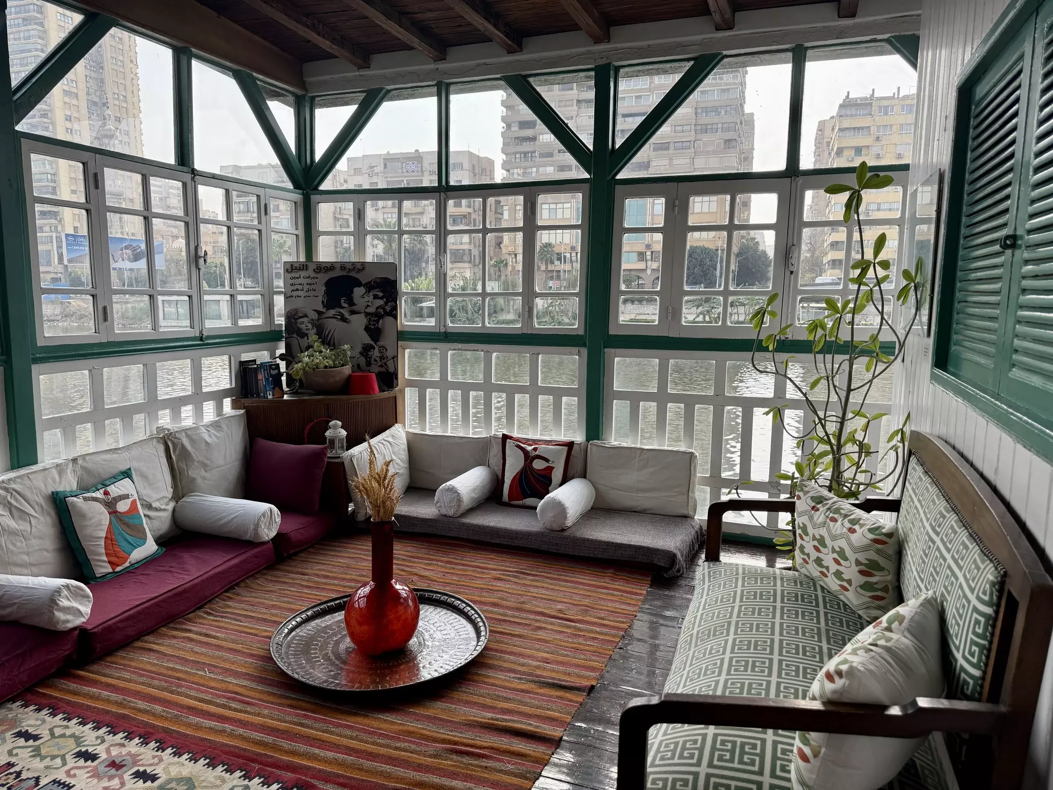 Gray and wine-colored floor sofas with bolster and accent pillows meet in the corner of a room with floor-to-ceiling windows and a multicolored striped rug. The windows look out on a river.