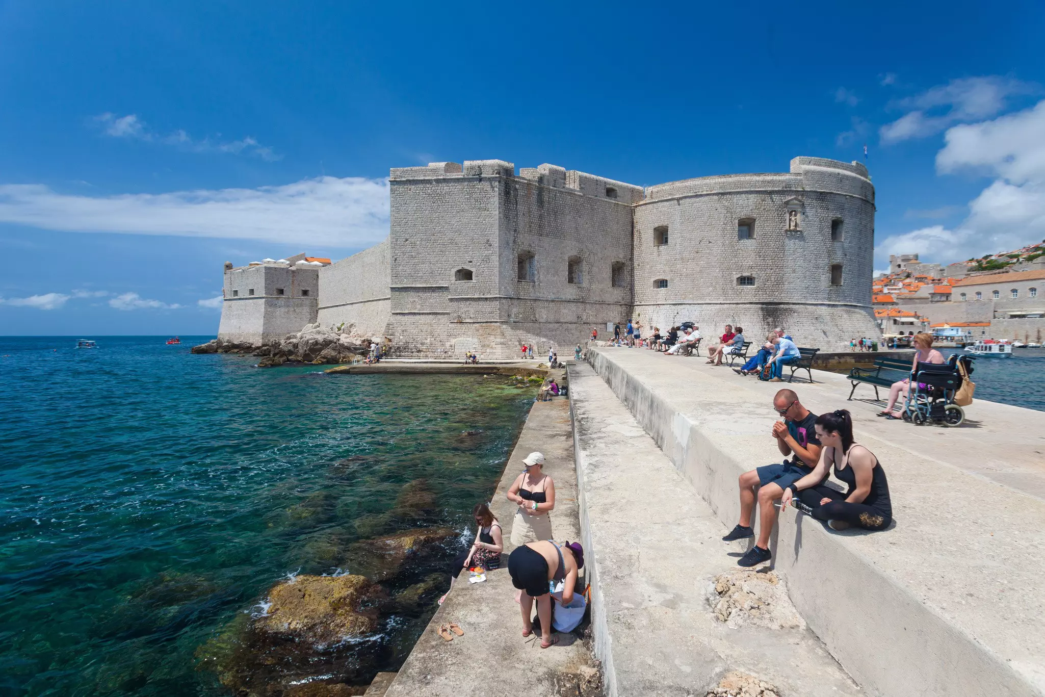Tourists sitting on bench and stairs on dock in front of the St. John fortress near the old port. Fortress houses the Maritime Museum and the aquarium.;