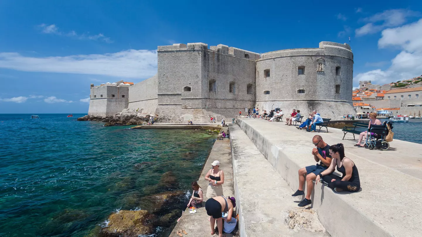 Tourists sitting on bench and stairs on dock in front of the St. John fortress near the old port. Fortress houses the Maritime Museum and the aquarium.;