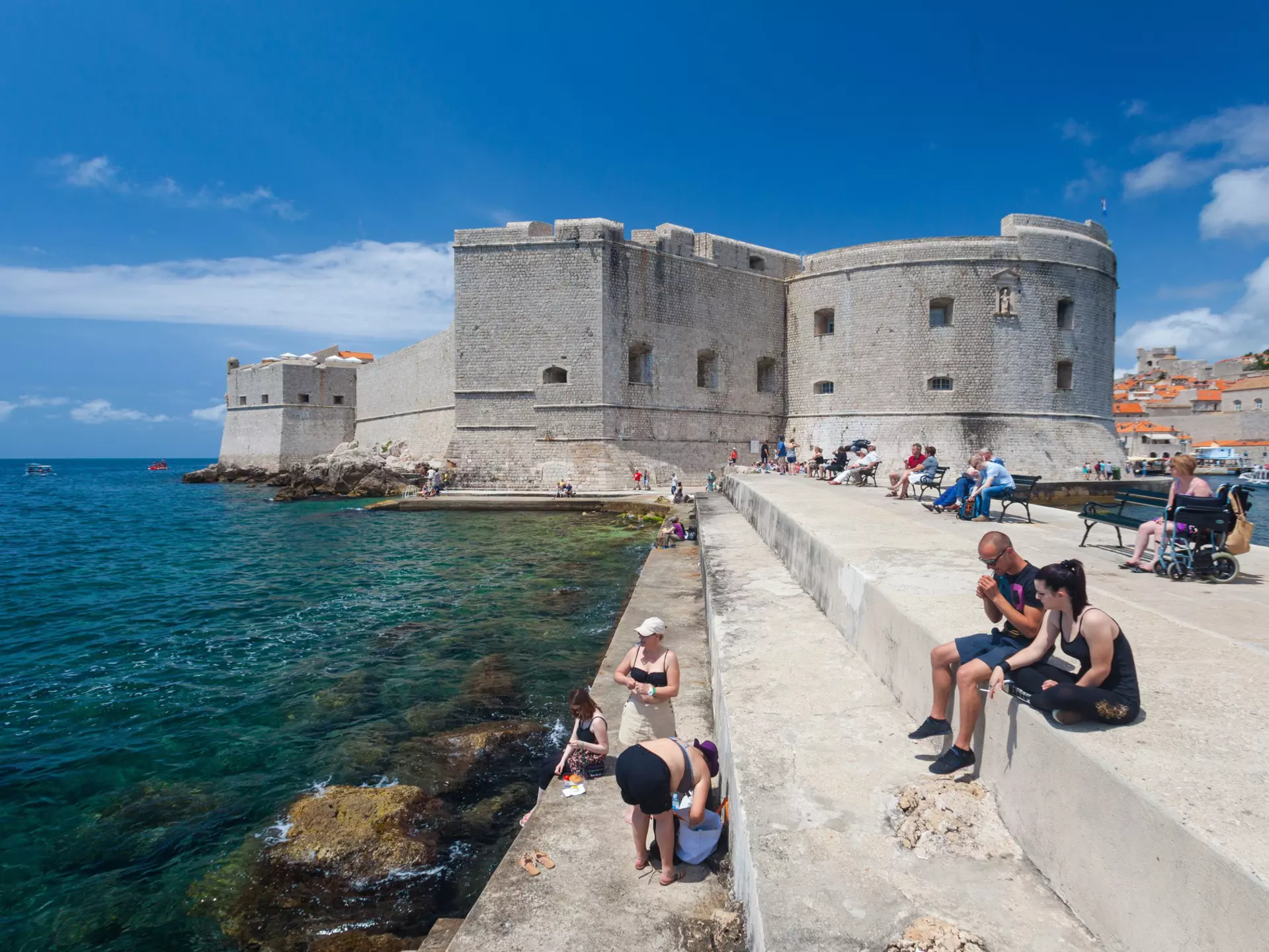 Tourists sitting on bench and stairs on dock in front of the St. John fortress near the old port. Fortress houses the Maritime Museum and the aquarium.;