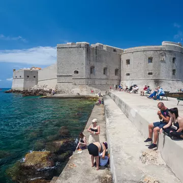 Tourists sitting on bench and stairs on dock in front of the St. John fortress near the old port. Fortress houses the Maritime Museum and the aquarium.;