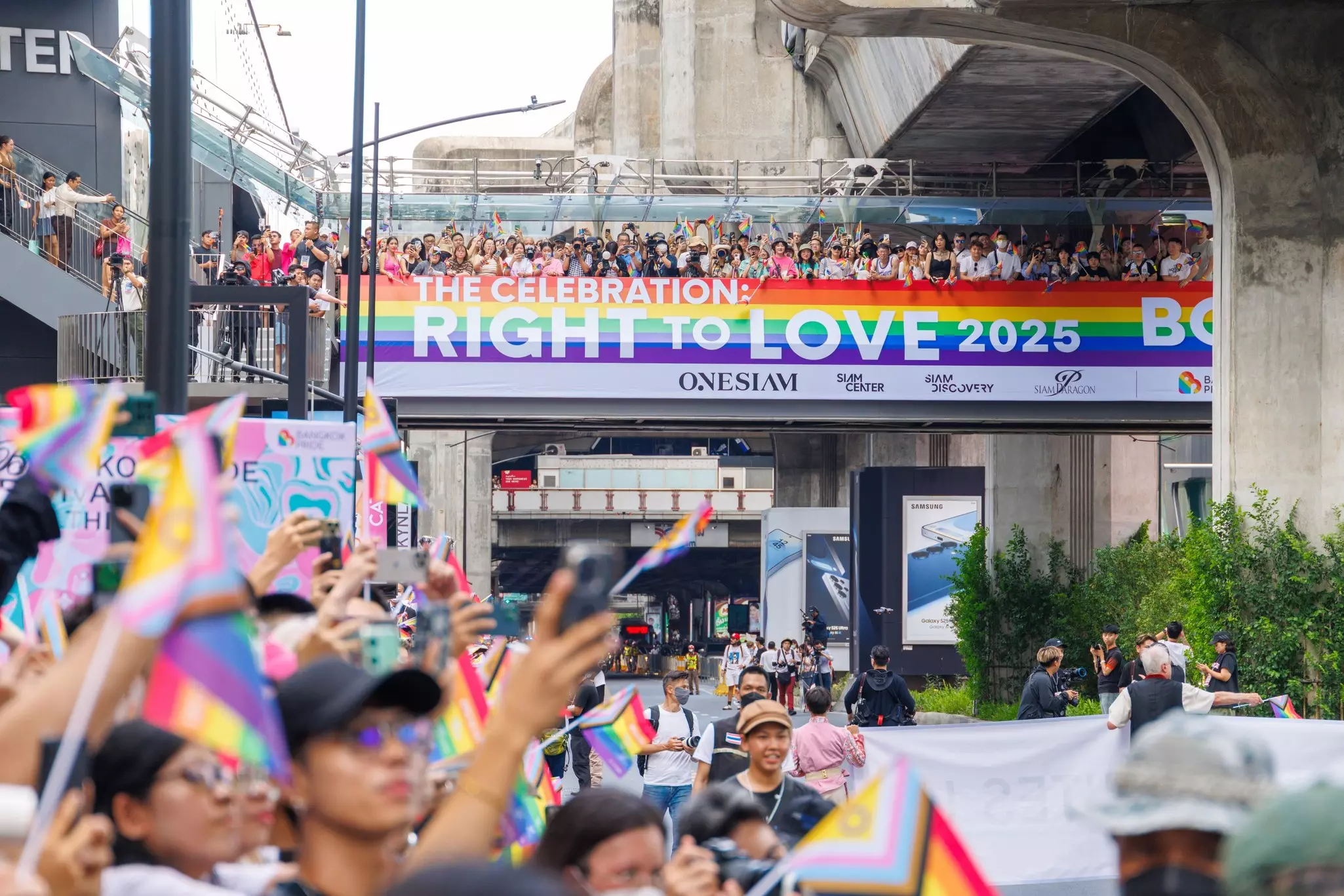 Crowds of people celebrate in the streets at an LGBT event, with rainbow flags and a large banner that says "The celebration: Right to Love 2025".