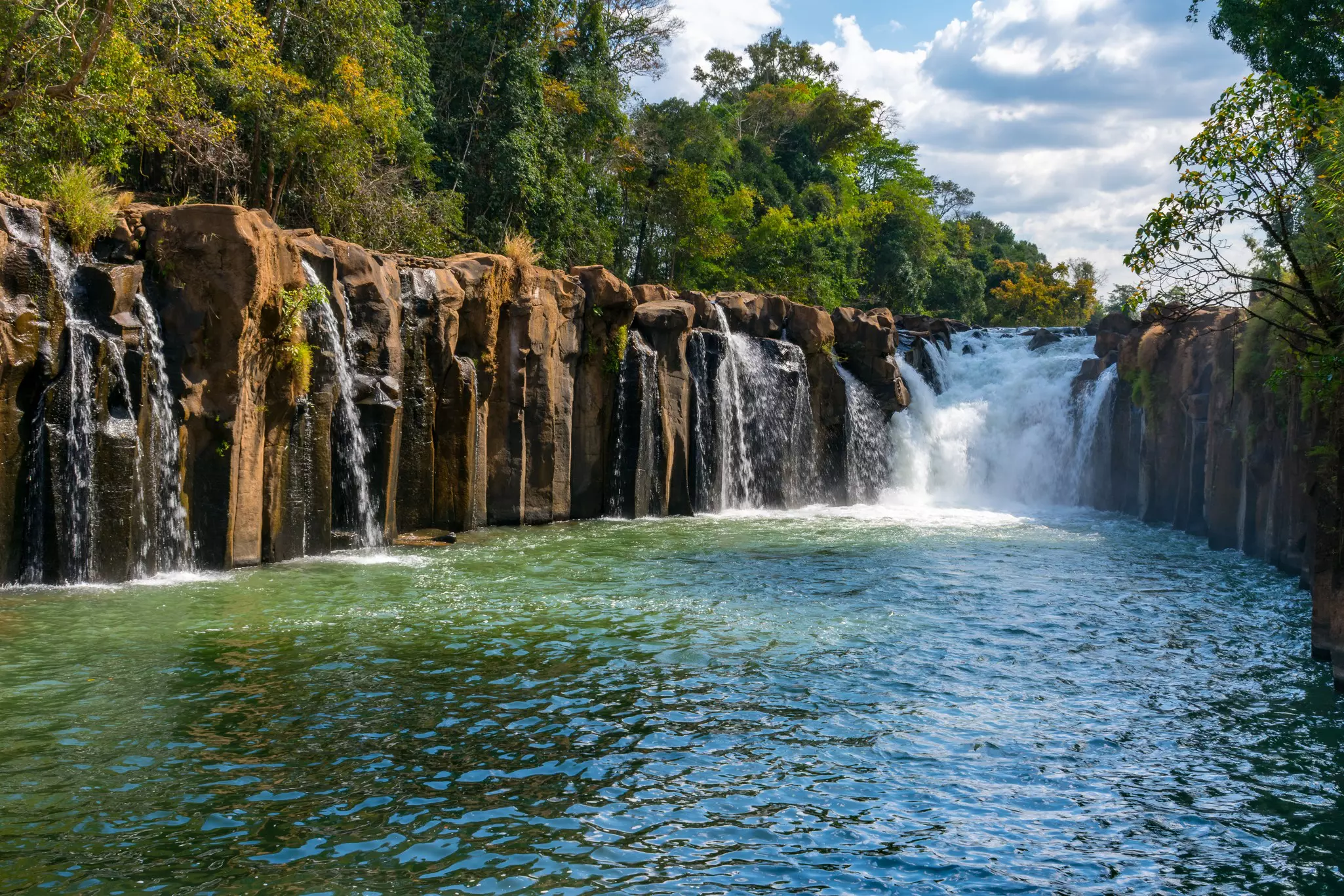 A waterfall cascades off rocks in jungle to land in a large pool.