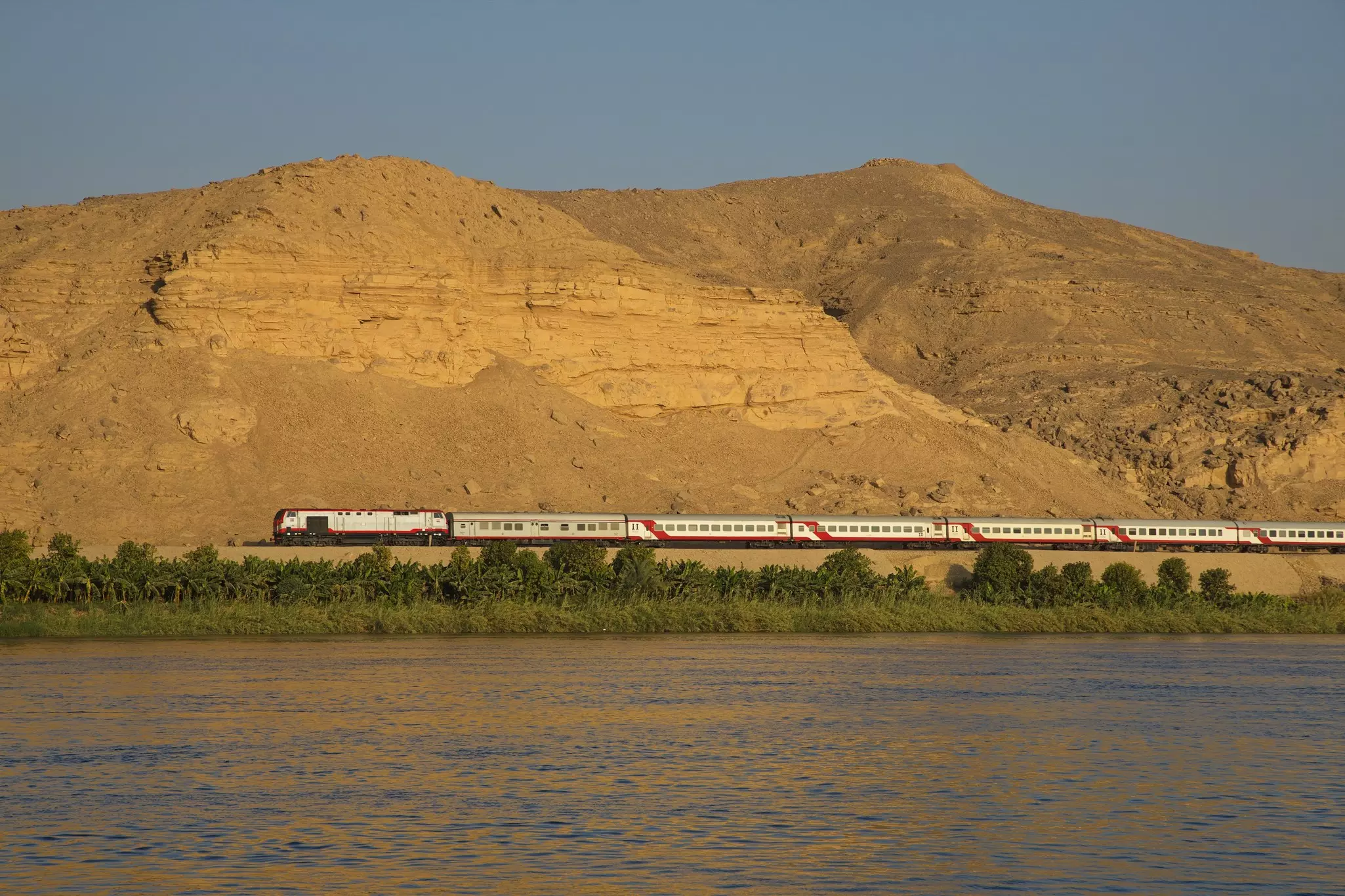 A train by a wide river passes in front of dry hills.