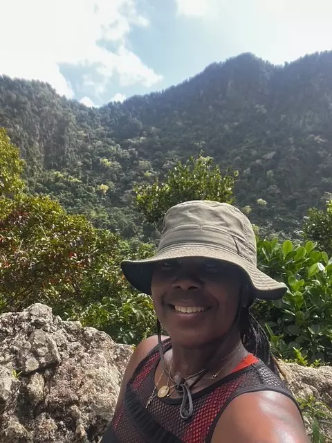 A woman wearing a green hat smiles at the camera as she pauses on a hike in a hilly region covered with dense foliage.