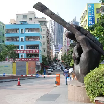 A large bronze hand holding a paintbrush on a plinth beside a street of pastel apartment blocks hung with Chinese art-supply signs.