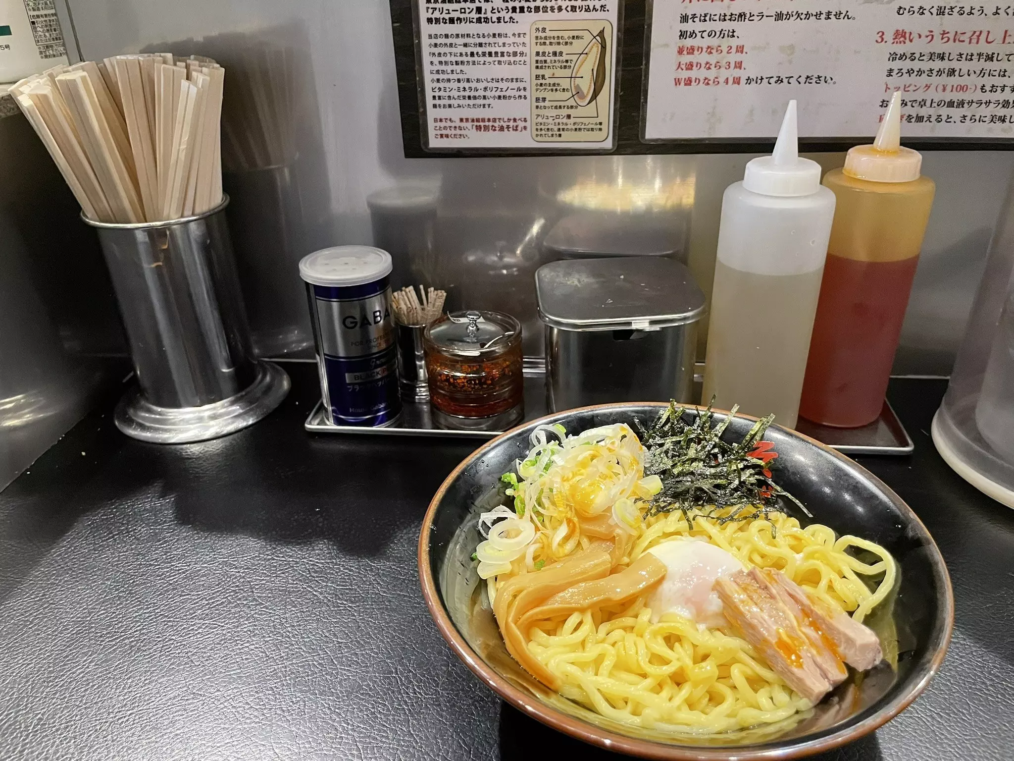 A bowl of ramen noodles on a dark counter with condiments and utensils.