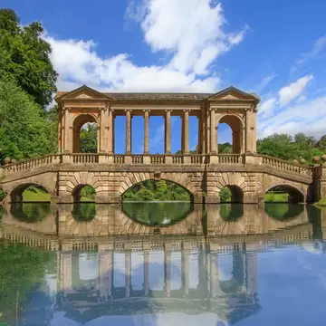 Colonnaded classical stone bridge mirrored in a still lake, framed by green woodland.