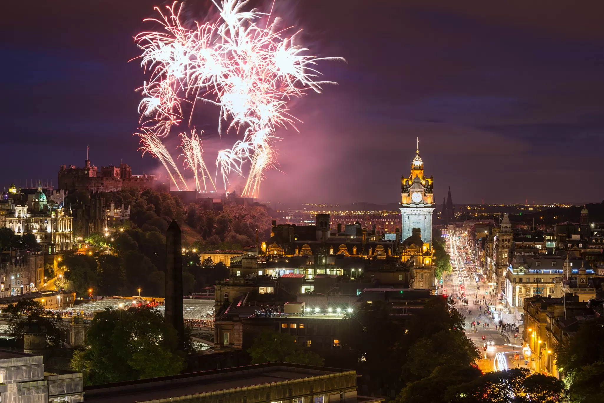 Fireworks lighting up the night sky with an illuminated clocktower and a busy street full of people