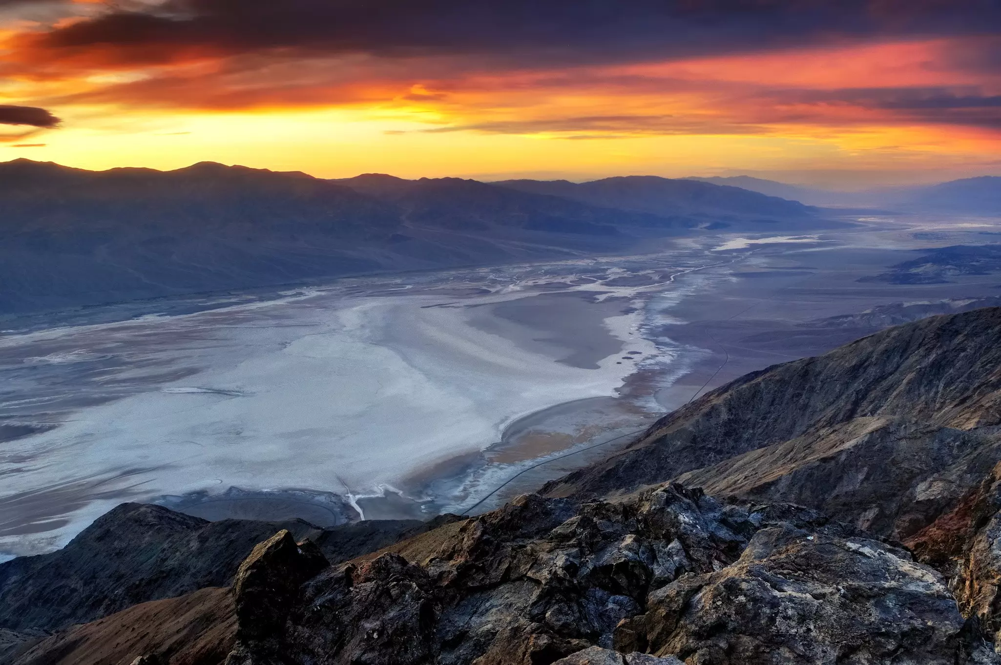 A valley of white salt flats surrounded by gray rocky hills and mountains. The sun is setting in the distance