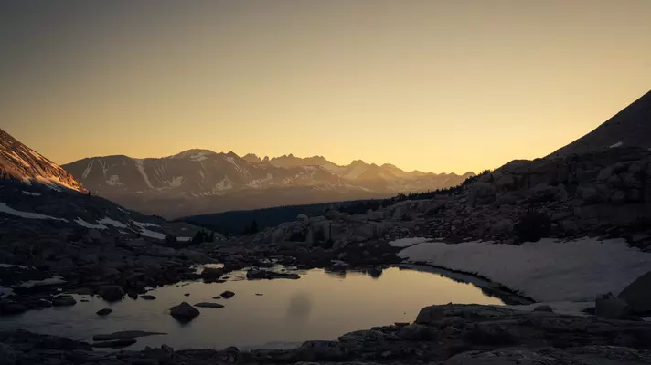 A mountain lake reflects the rising sun as the sky lights up in pale yellow.