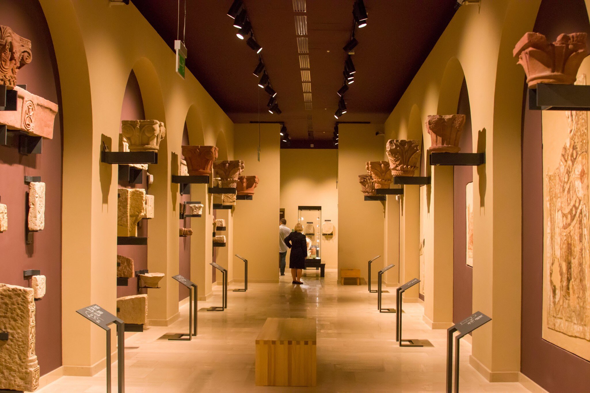 Carved stone capitals on wall brackets line an arched ochre hall where two visitors study a display case.