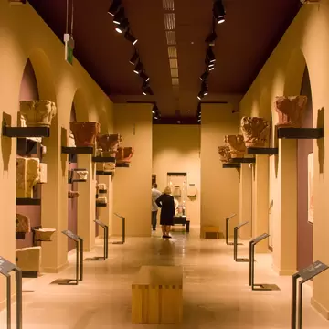 Carved stone capitals on wall brackets line an arched ochre hall where two visitors study a display case.