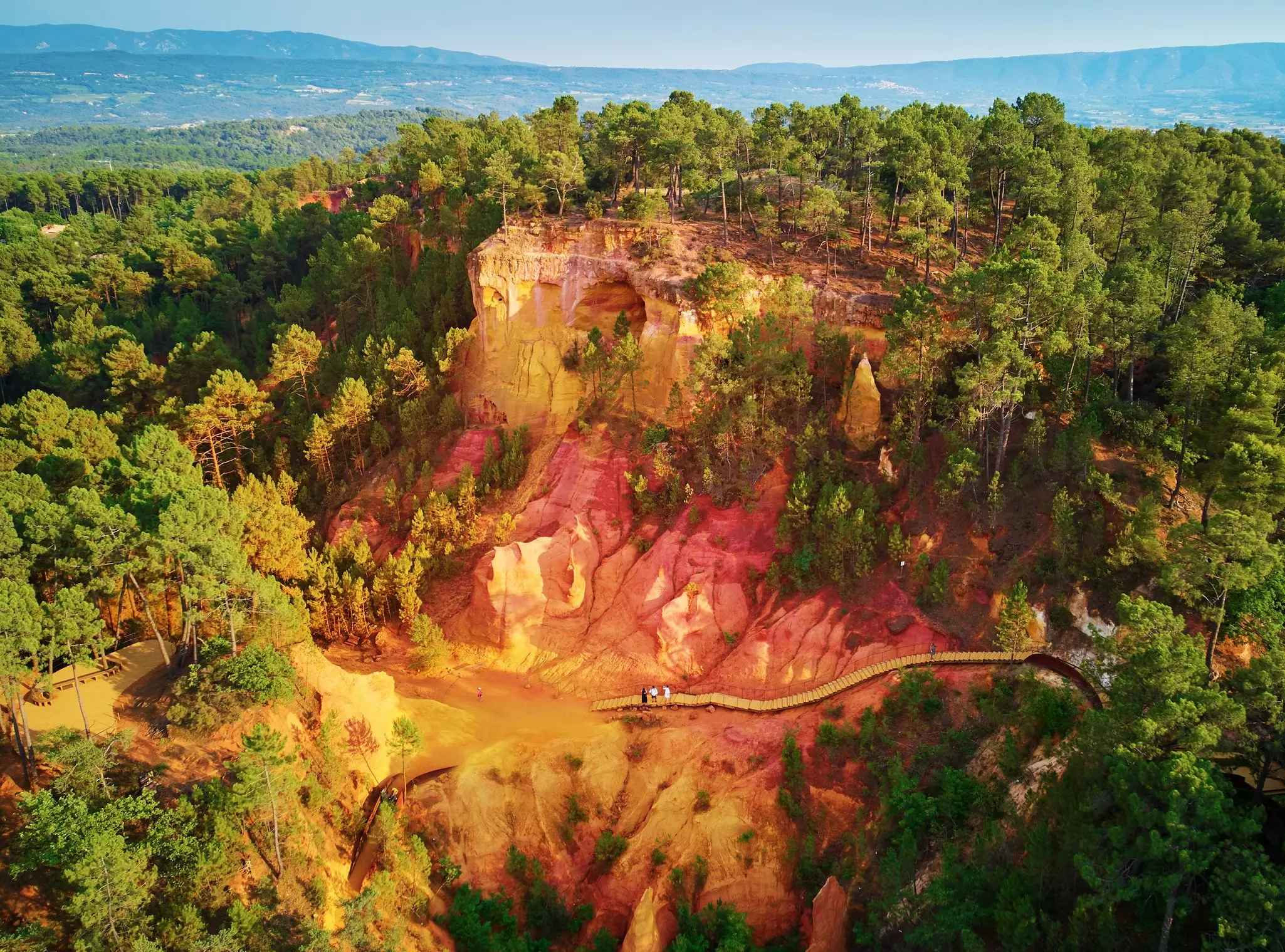 A boardwalk through an orange and red sandy landscape with sheer cliffs covered in woodland.