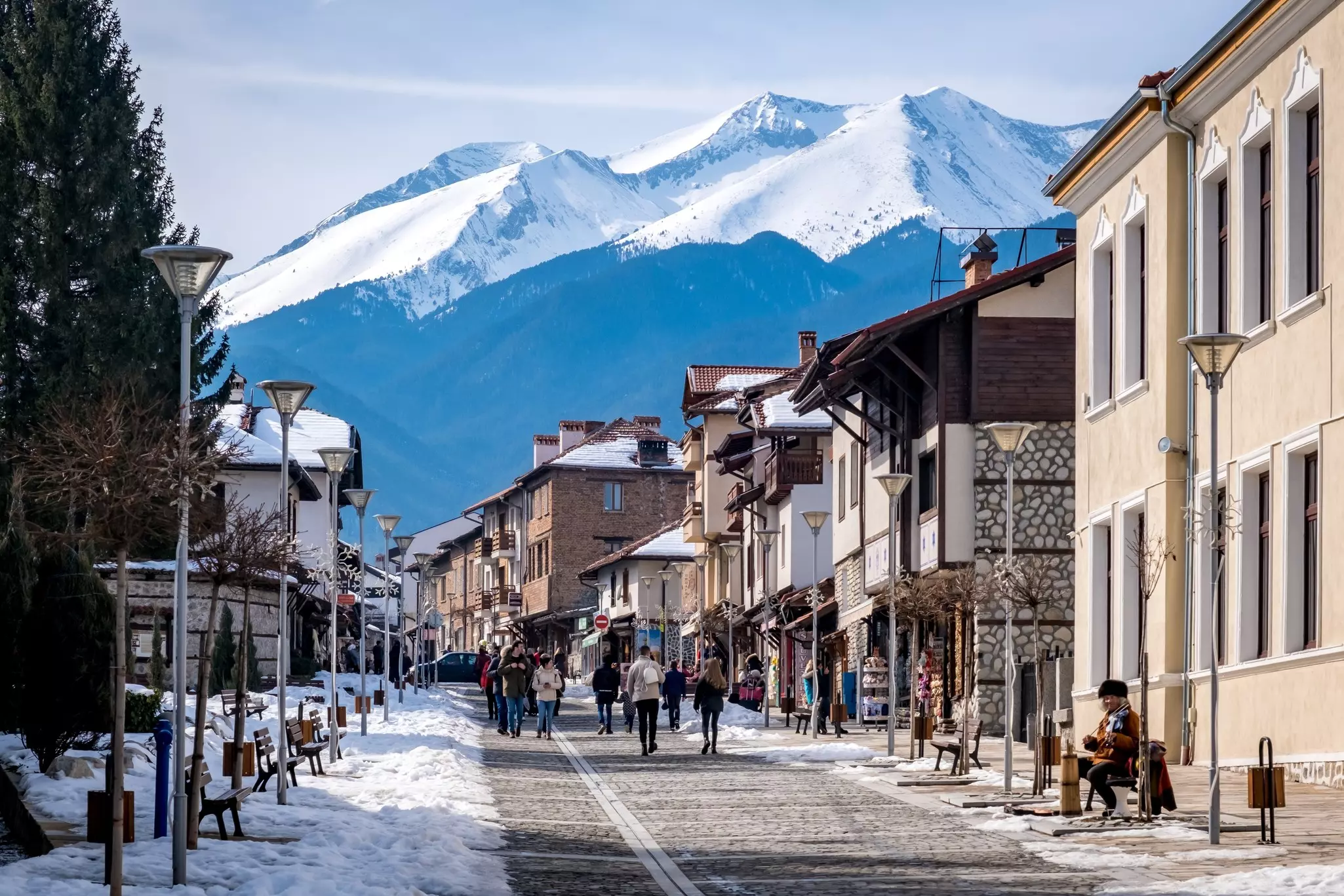 People walk on a cobblestone street through a town in Bulgaria; there is snow on the roofs and to the side of the roadway. A tall snowcapped peak looms over the town in the background.