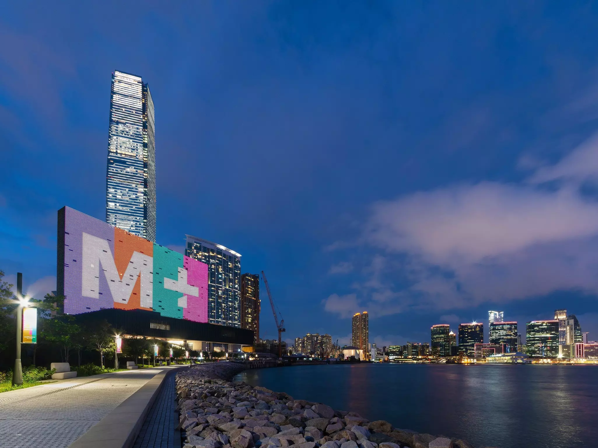 Evening view of harborside prominade with large modern M+ Museum and city skyline in the distance.