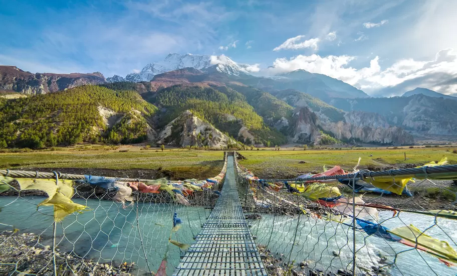 Nepal: steel suspension bridge hung with prayer flags over a glacial river, snowcapped Annapurna peak behind