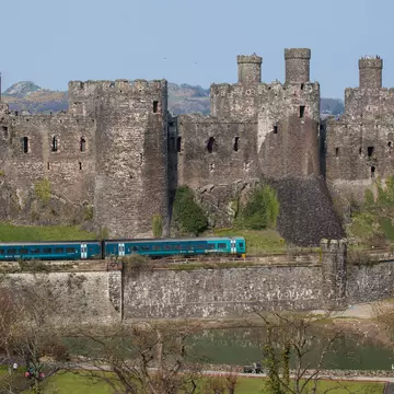 Comwy, Wales - April 9, 2015: Conwy Castle and a Passenger Train North Wales. The railway lone runs along the base of the medeval castle and the Virgin Trains service passes the castle. People can be seen on the catsle walls. The sky is clear and blue.
472088294
Surrounding Wall, Single Line, Travel, Service, People Traveling, Public Transportation, High Speed Train, Train Tunnel, Non-Urban Scene, Copy Space, Voyager, Retaining Wall, Conwy Castle, Conwy, Coastline, Caernarfon, Stone Material, Covering, Fortified Wall, Business Travel, Medieval, Blue, In A Row, Welsh Culture, Cultures, Industry, Travel Destinations, Transportation, Tourist, People, Wales, UK, Tree, Mountain Pass, Sky, Sea, Wall, Railroad Track, Castle, Passenger Train, Train, Mode of Transport, Embankment