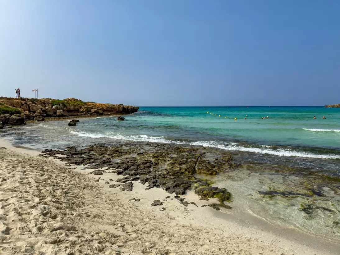 Pale sand meets dark rocks and shallow turquoise sea, with swimmers near a line of buoys and a rocky headland at left.