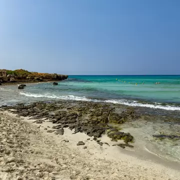 Pale sand meets dark rocks and shallow turquoise sea, with swimmers near a line of buoys and a rocky headland at left.