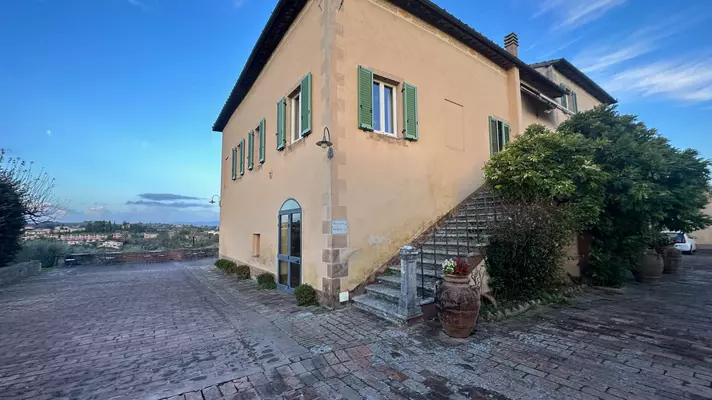 A staircase leads up the side of a tan building with green shutters. A cobblestone patio surrounds the building, and there are distant views of the countryside.