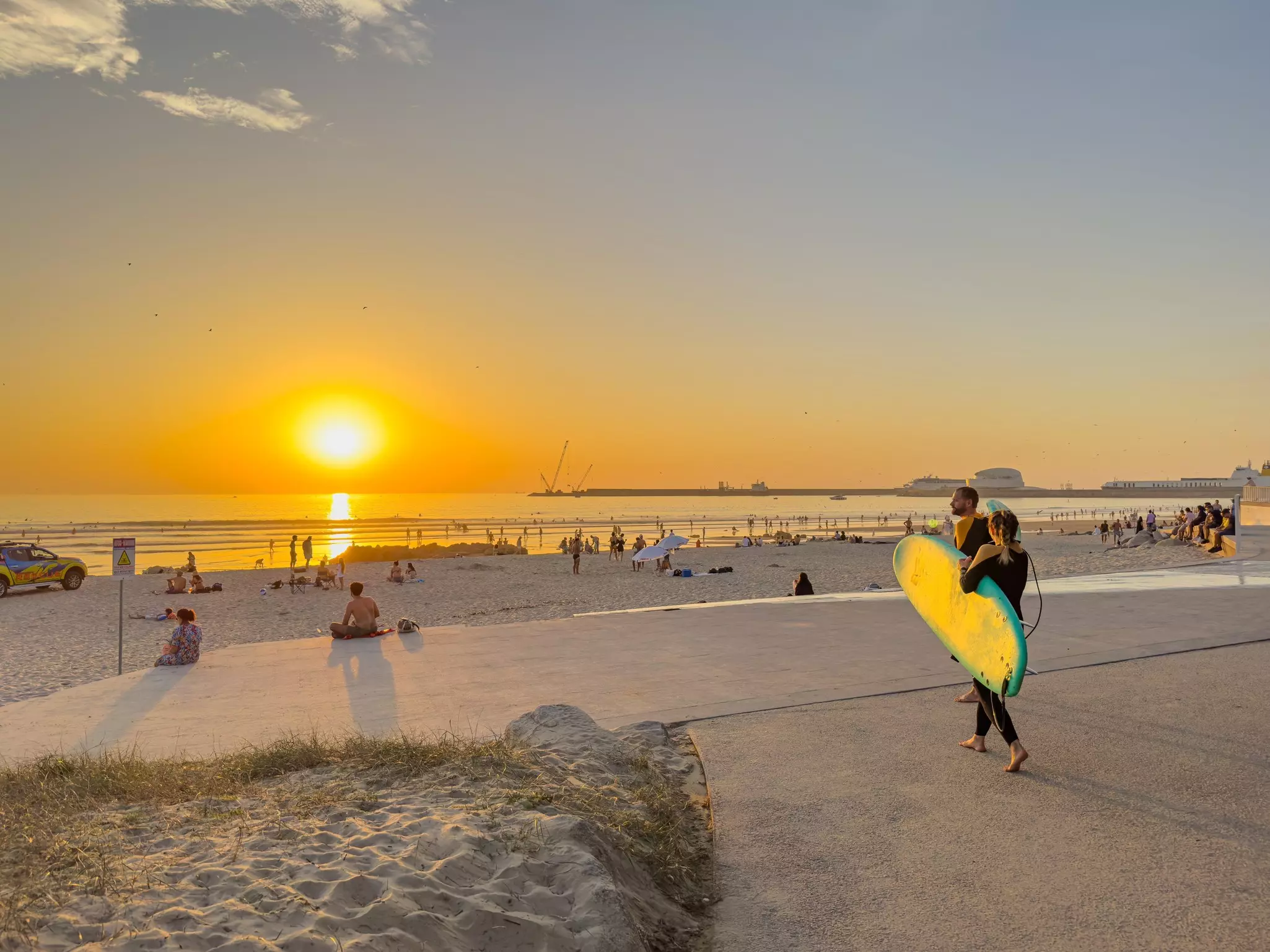 A surfer carries a surfboard toward a beach with people on the sand near Porto, Portugal, at sunset.