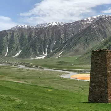 A ruined stone tower stands on green meadow above a braided river with orange mineral deposits below snowy peaks.