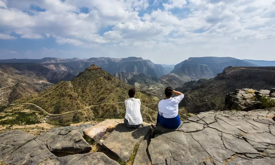 Two women sitting on a rock ledge overlooking deep canyons and terraced slopes of the Jebel Akhdar mountains, Oman
