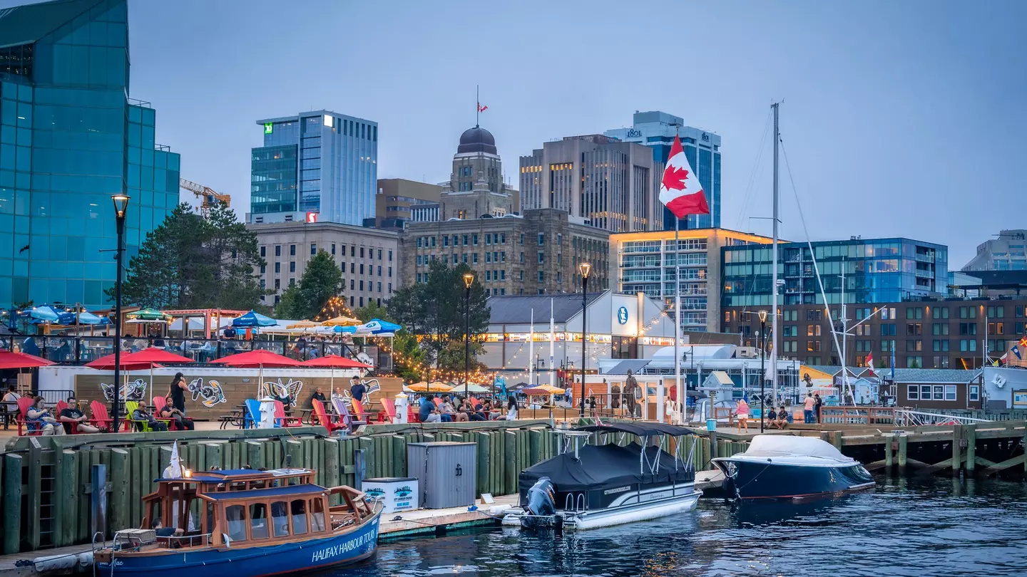 A city harbor. People wander along the waterfront and three small boats are docked nearby.