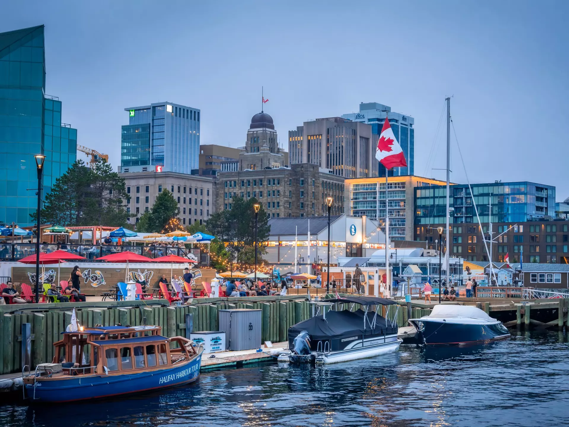 A city harbor. People wander along the waterfront and three small boats are docked nearby.