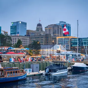 A city harbor. People wander along the waterfront and three small boats are docked nearby.