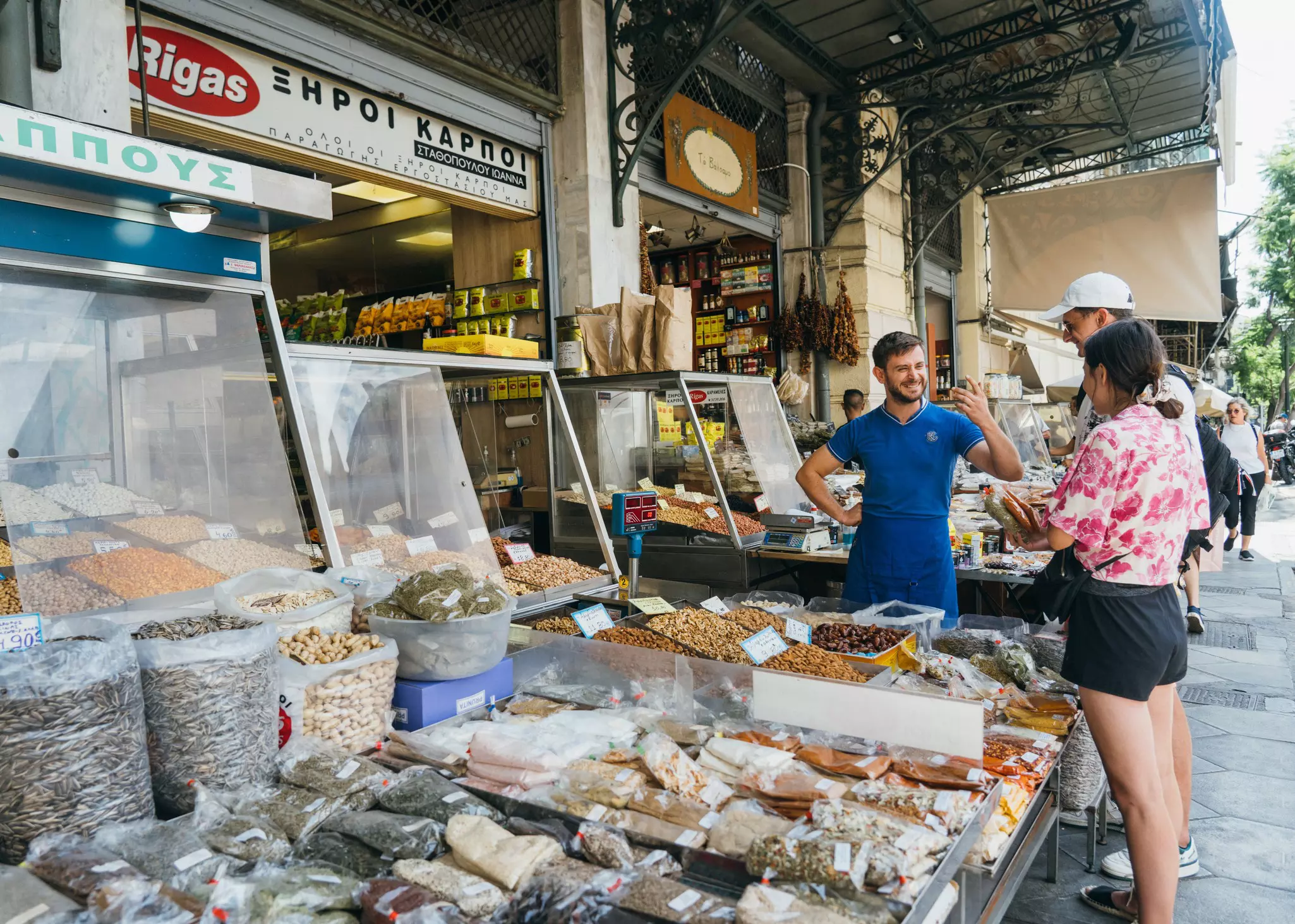 A market vendor chats with two customers in front of a display of nuts and seeds.